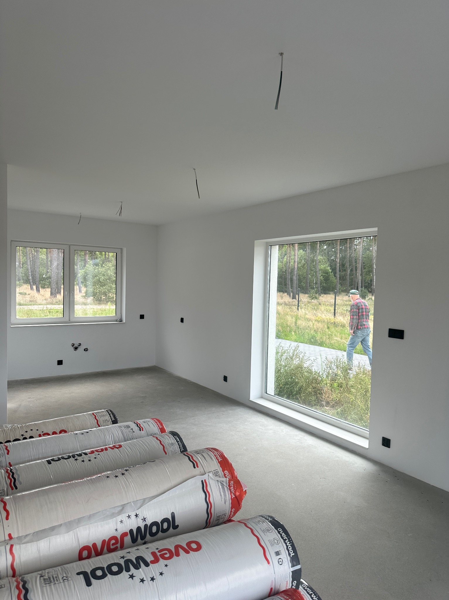 Surowe wnętrze z oknami i rolkami izolacji Overwool na podłodze. W oknie widoczna osoba. W tle las. Białe ściany z wystającymi kablami.