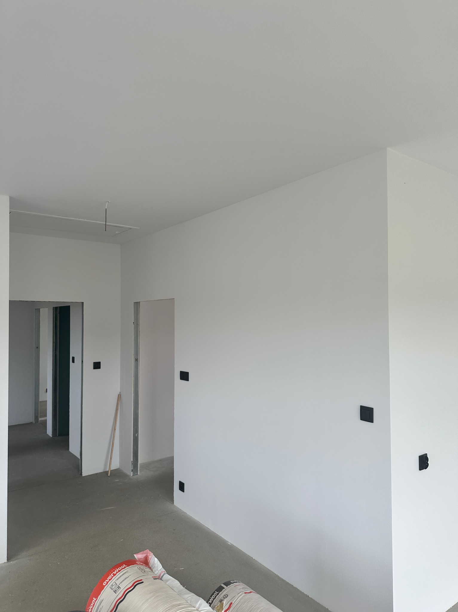 Wnętrze w trakcie remontu: białe ściany, surowa podłoga, widoczne puszki elektryczne. W tle otwarte przejścia do innych pomieszczeń. Na podłodze leżą worki z materiałem izolacyjnym.