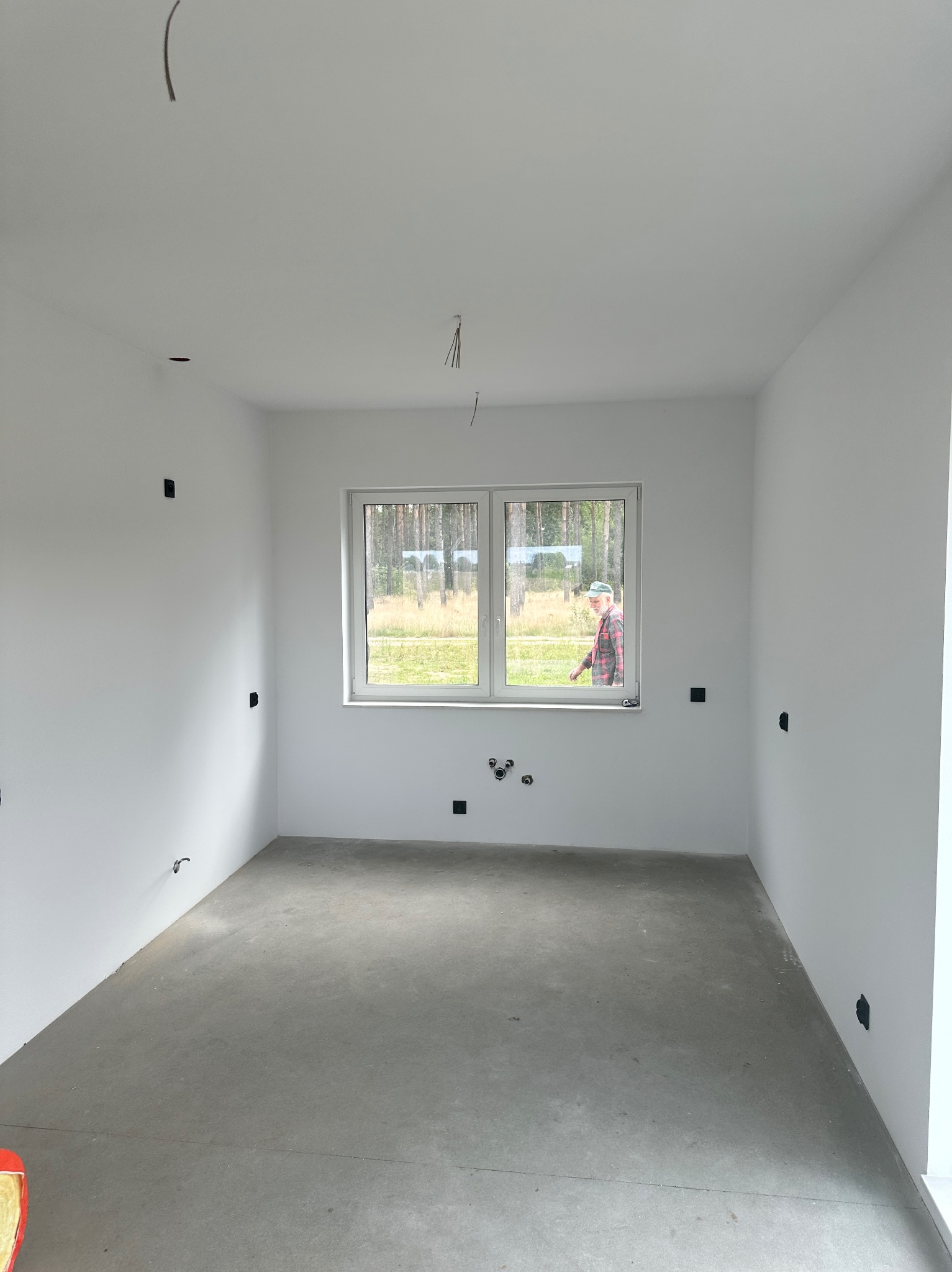 Surowe wnętrze pokoju z białymi ścianami, szarą podłogą i oknem. Widoczne wystające kable i puszki elektryczne. Osoba w oknie w tle. Stan deweloperski przed wykończeniem.