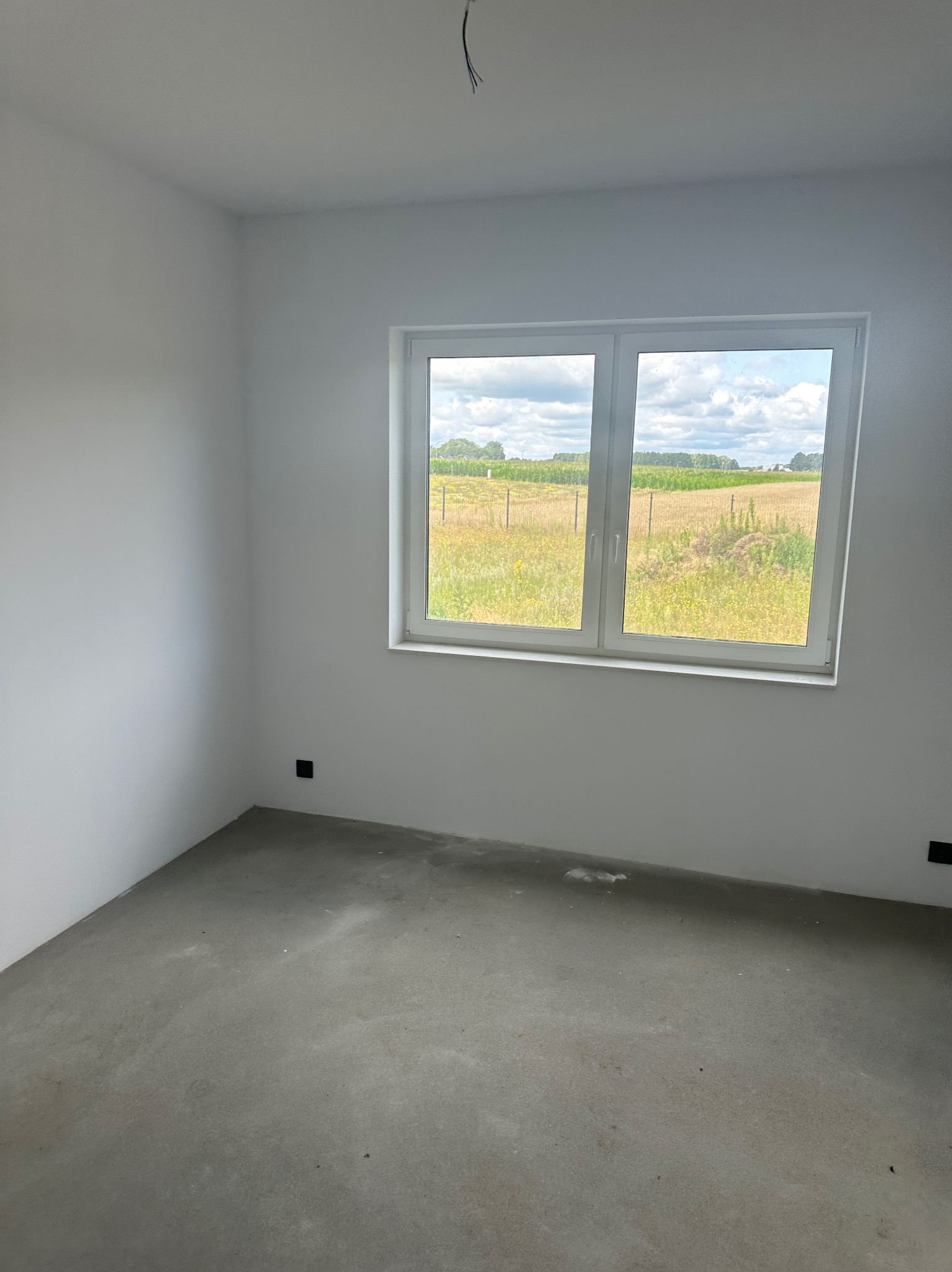 Pusty, jasny pokój z surową podłogą i białymi ścianami. Okno z widokiem na pole. W tle widoczne drzewa i niebo. W ścianach widoczne gniazdka elektryczne.