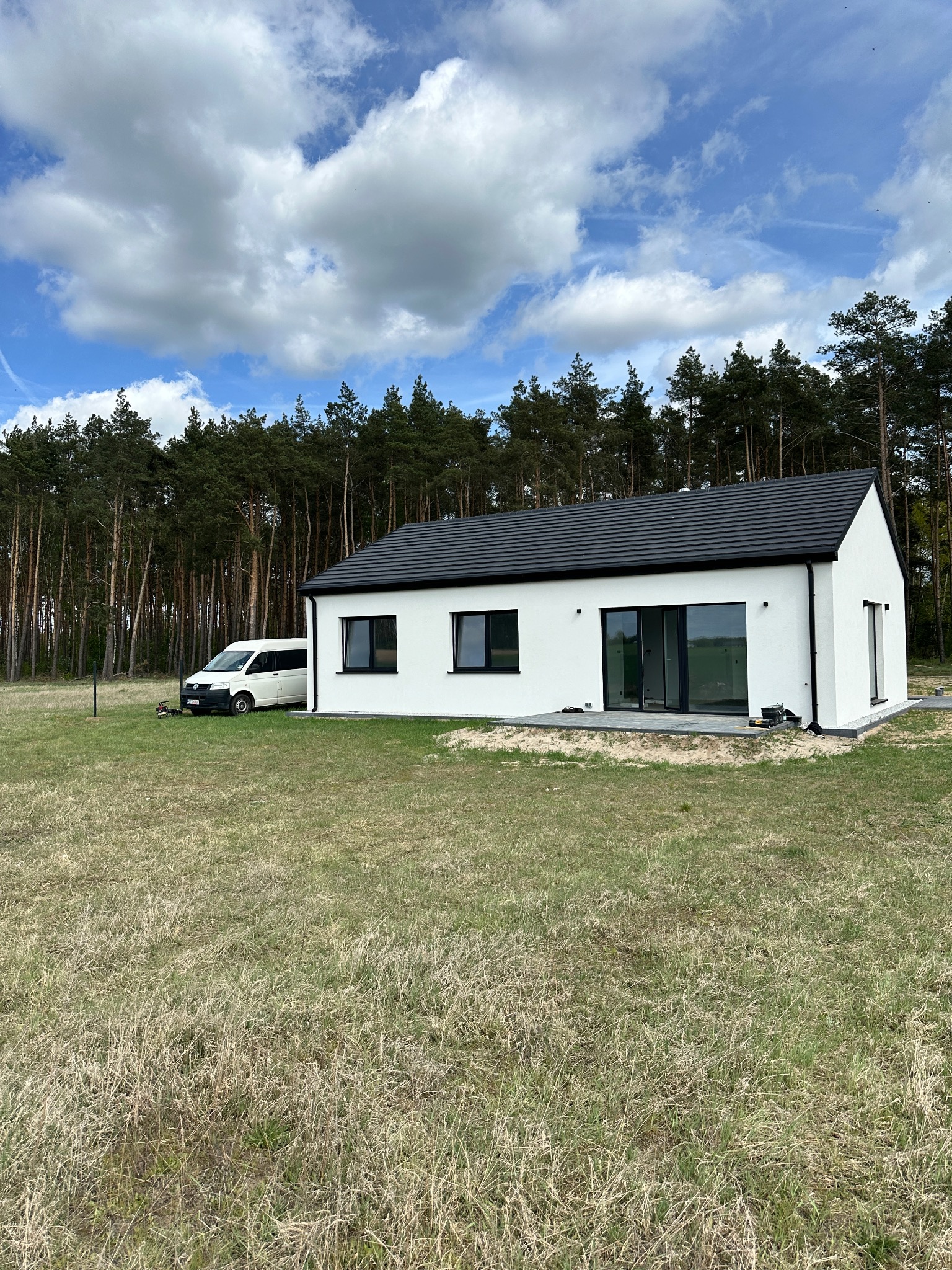 Biały, parterowy dom z czarnym dachem na tle lasu i błękitnego nieba z chmurami. Na trawie stoi biały bus. Widoczne okna i taras z szarych płyt.
