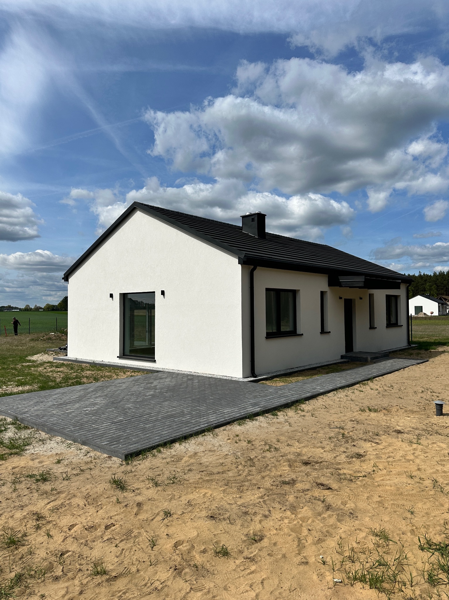 Nowoczesny, parterowy dom z białymi ścianami i czarnym dachem na działce z kostką brukową. Widoczne okna z czarnymi ramami i fragment zielonego krajobrazu w tle.