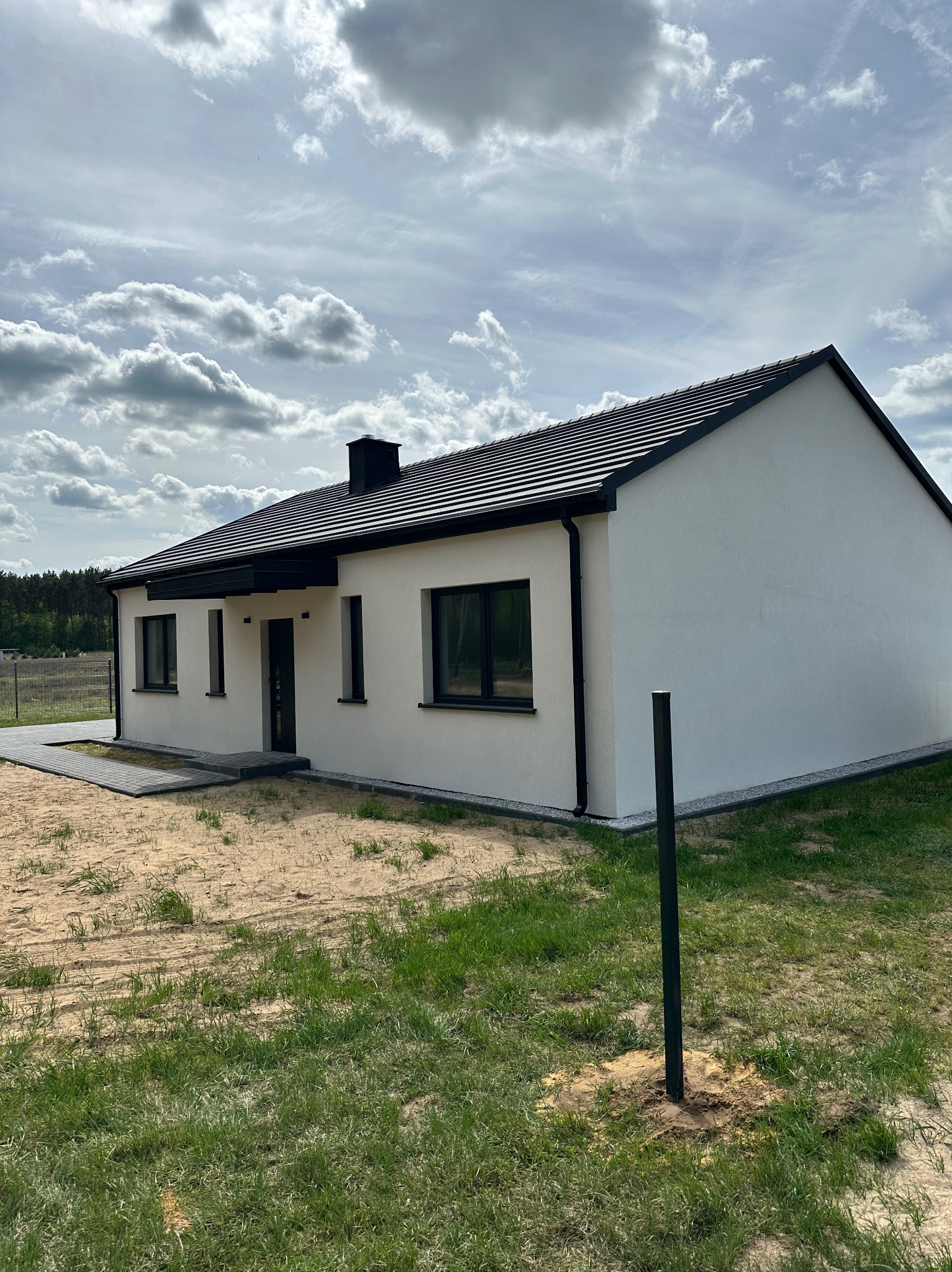 Biały, parterowy dom z czarną dachówką i stolarką okienną na działce. Widoczne fragmenty trawnika i nieba z chmurami. Minimalistyczna architektura.