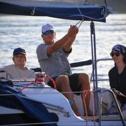 Lipińskifoto - Rodzinny rejs żaglówką: nastolatek obsługuje liny, młodszy chłopiec siedzi obok, a dziewczyna w bandanie uśmiecha się w tle, morze i słońce.