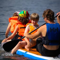 Lipińskifoto - Rodzinna wyprawa na desce SUP: dzieci w kamizelkach płyną po jeziorze, wiosłując pod okiem instruktora. Aktywny wypoczynek na wodzie w słoneczny dzień.