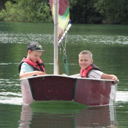 Lipińskifoto - Dwóch chłopców w kamizelkach płynie łódką po jeziorze w Częstochowie. Uśmiechnięte dzieci na tle zieleni i wody, żaglówka z kolorowym żaglem.