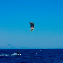 Kitesurfer płynie po ciemnoniebieskim morzu pod błękitnym niebem, w tle zarys gór. Widoczny kolorowy latawiec i ślad wody za deską.