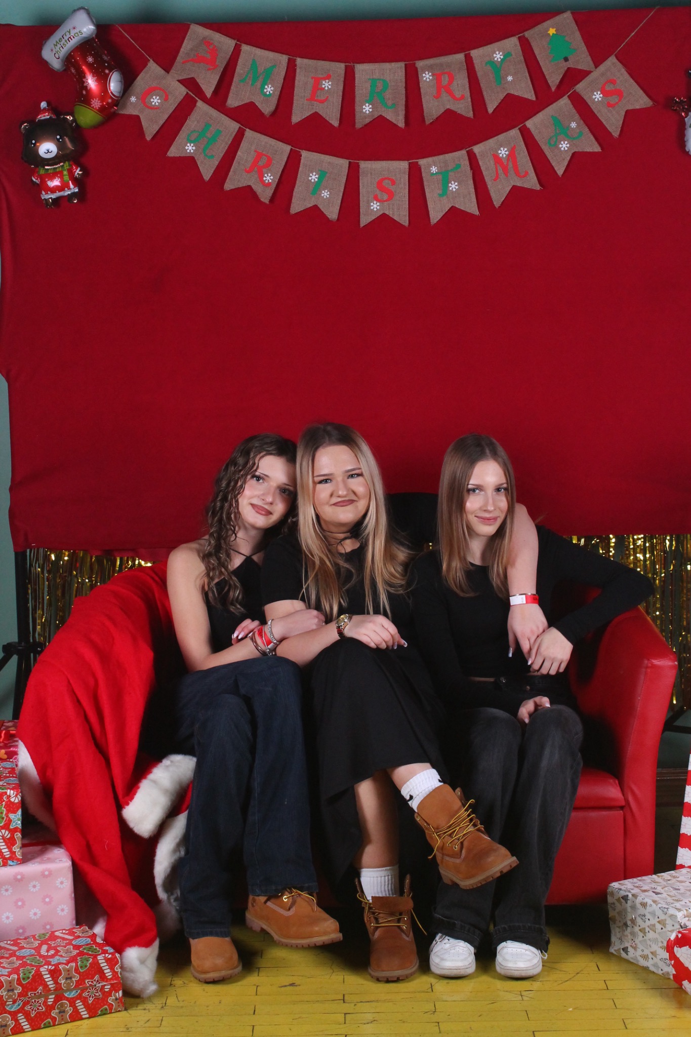Trzy nastolatki siedzą na czerwonej kanapie na tle świątecznej dekoracji z napisem 'Merry Christmas'. Prezentowe pudełka i czapka Mikołaja dodają klimatu.