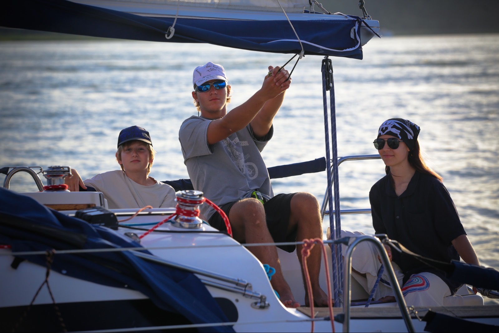 Rodzinny rejs żaglówką: nastolatek obsługuje liny, młodszy chłopiec siedzi obok, a dziewczyna w bandanie uśmiecha się w tle, morze i słońce.