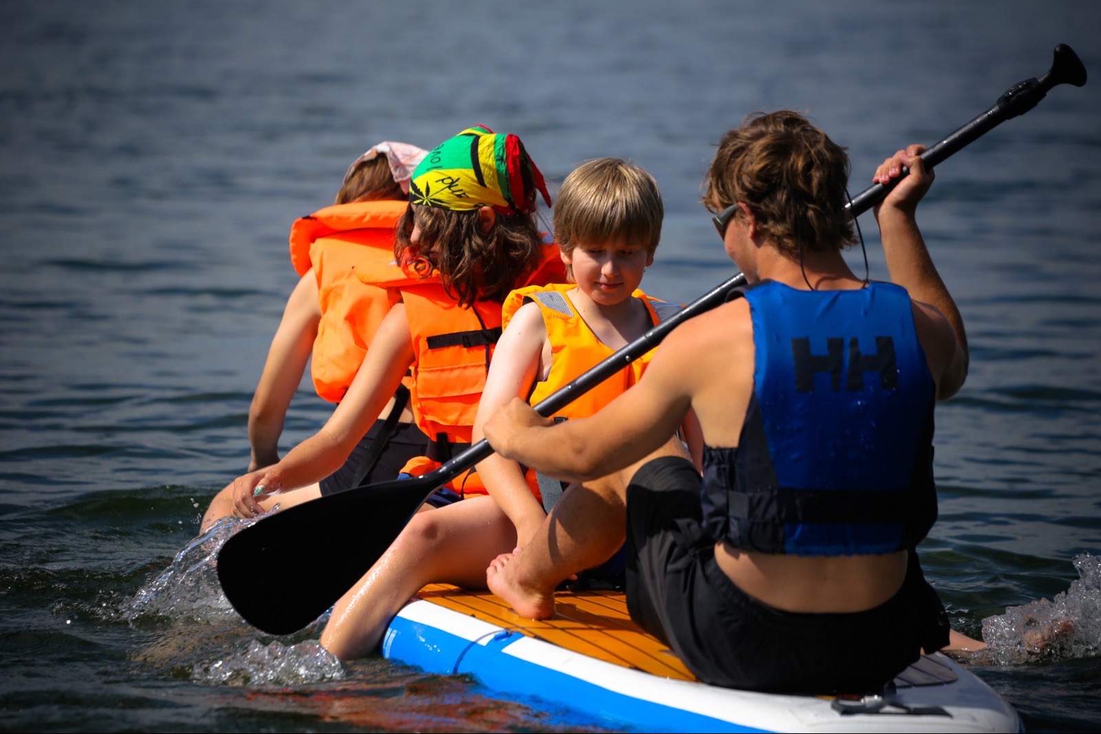 Rodzinna wyprawa na desce SUP: dzieci w kamizelkach płyną po jeziorze, wiosłując pod okiem instruktora. Aktywny wypoczynek na wodzie w słoneczny dzień.