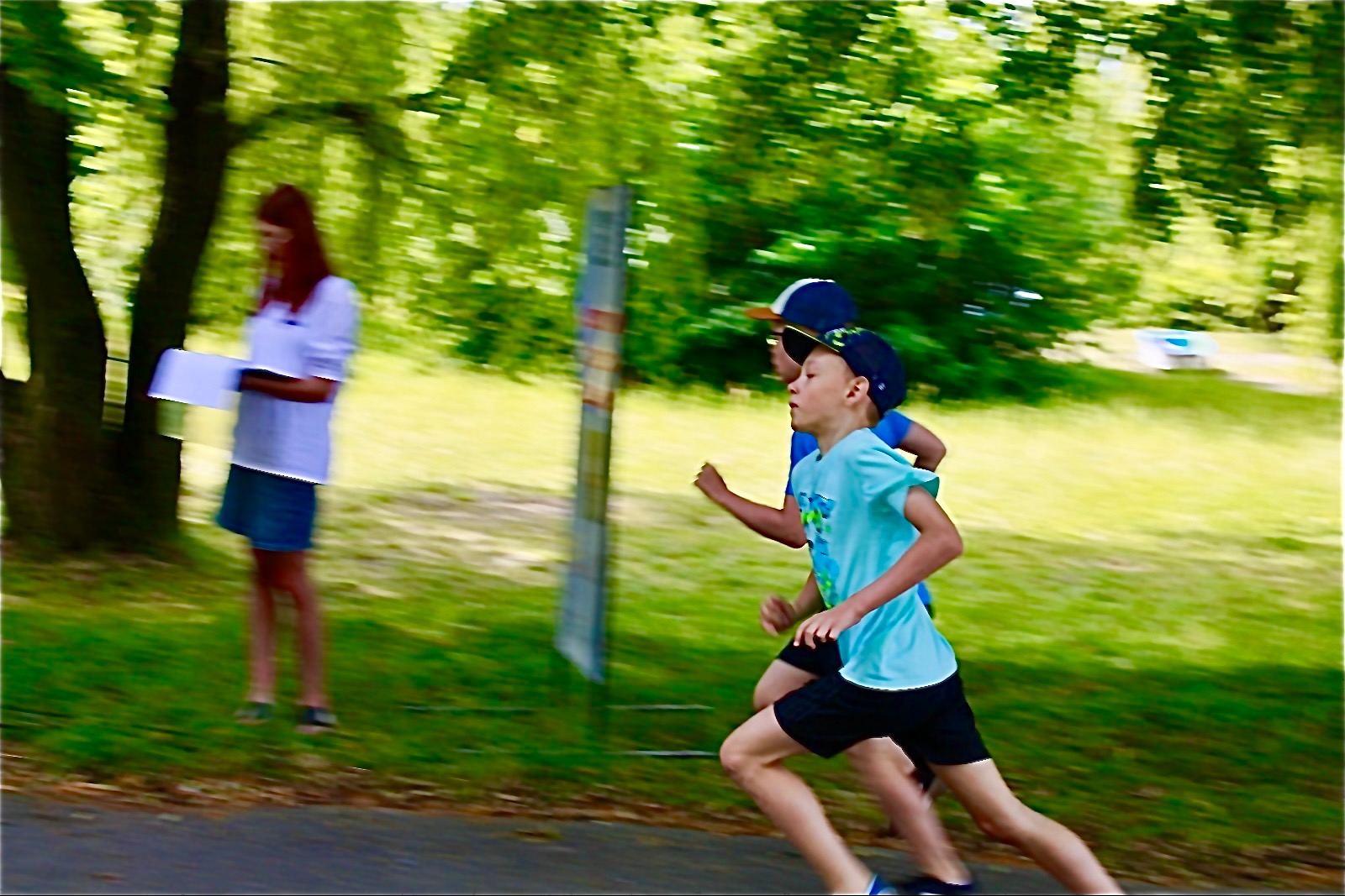 Dwóch chłopców w biegu podczas zawodów sportowych na świeżym powietrzu, sędzia z listą wyników w tle. Dynamiczne ujęcie, zieleń drzew i trawy.