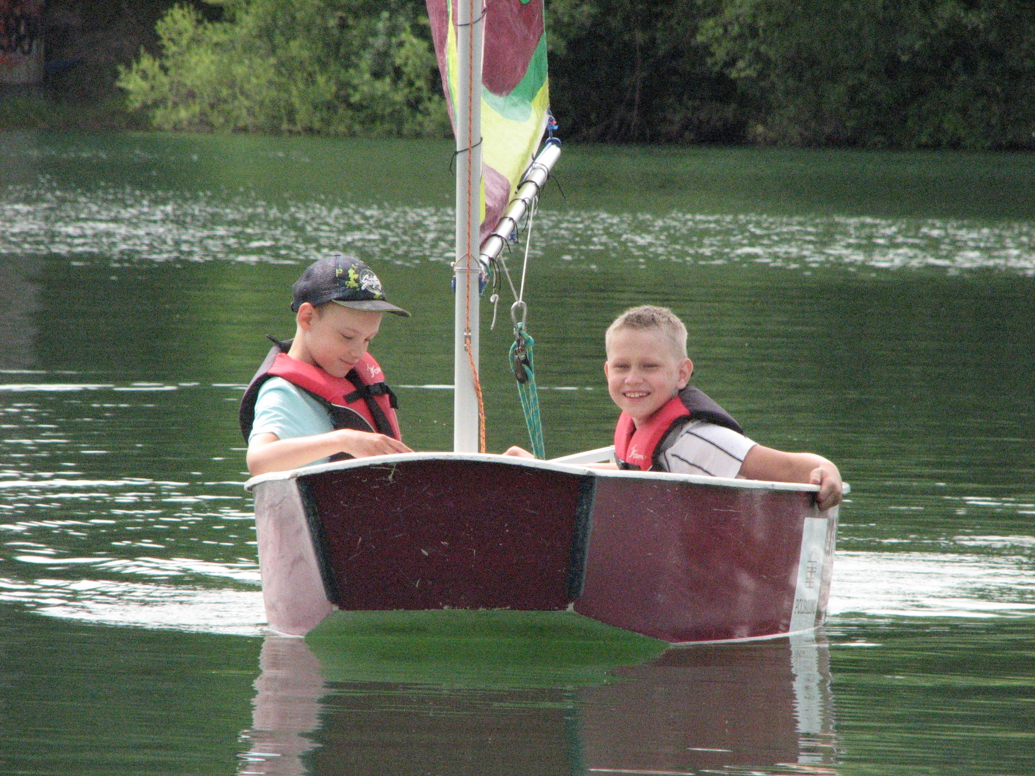 Dwóch chłopców w kamizelkach płynie łódką po jeziorze w Częstochowie. Uśmiechnięte dzieci na tle zieleni i wody, żaglówka z kolorowym żaglem.