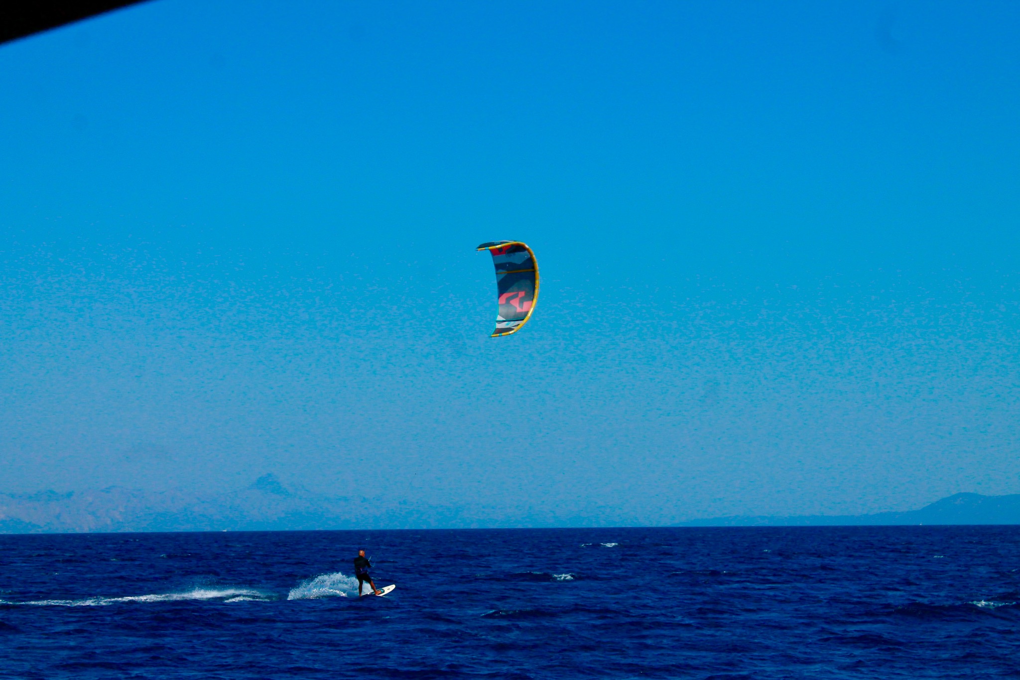Kitesurfer płynie po ciemnoniebieskim morzu pod błękitnym niebem, w tle zarys gór. Widoczny kolorowy latawiec i ślad wody za deską.