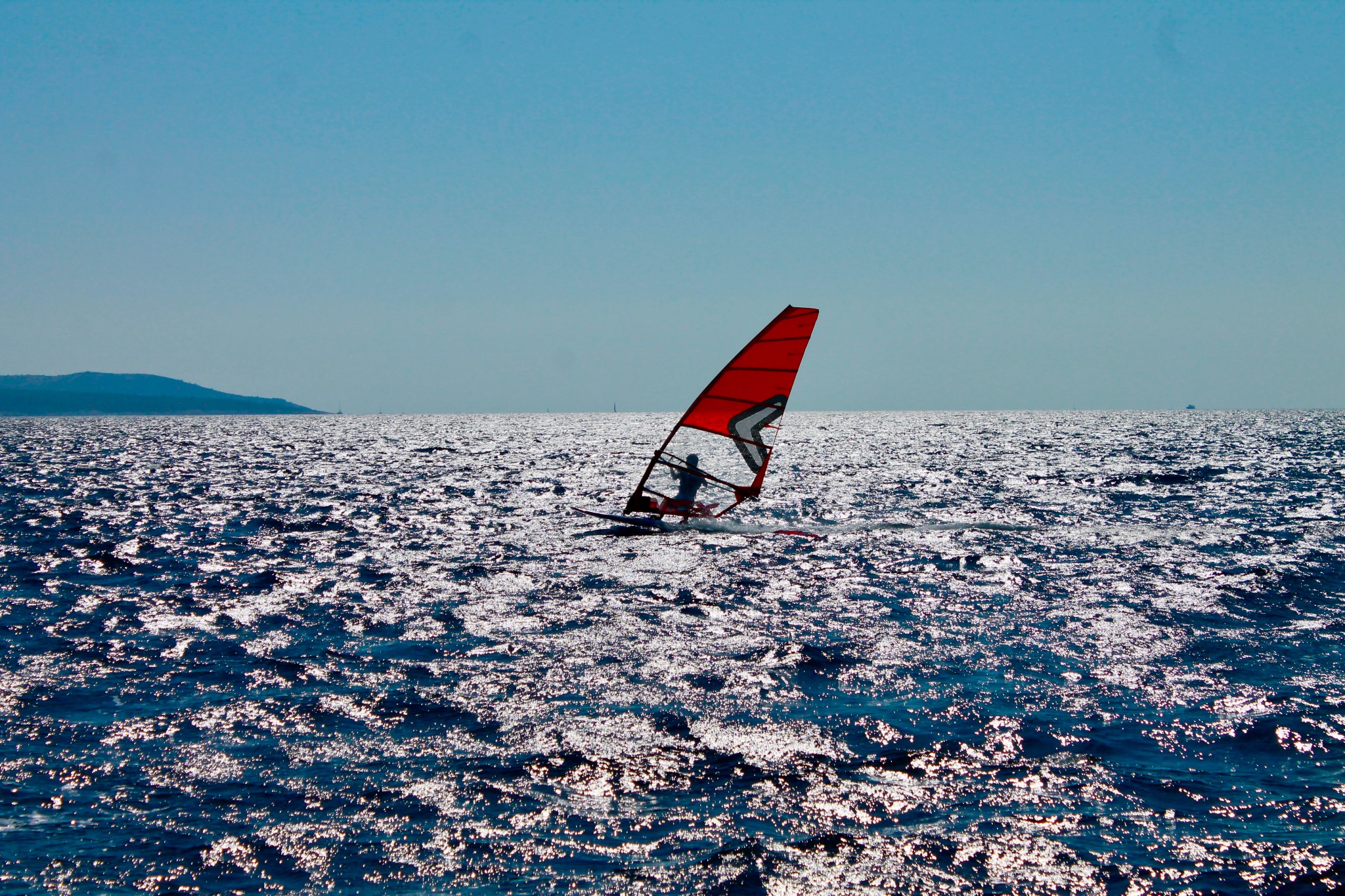 Windsurfer z czerwoną żaglą na połyskującej wodzie w słoneczny dzień. W tle widoczny zarys lądu.