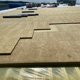 Daniel-Bud Grobelny Daniel - Układanie izolacji z wełny mineralnej na płaskim dachu budynku przemysłowego w słoneczny dzień. Widoczne rolki papy i pracownicy w oddali.