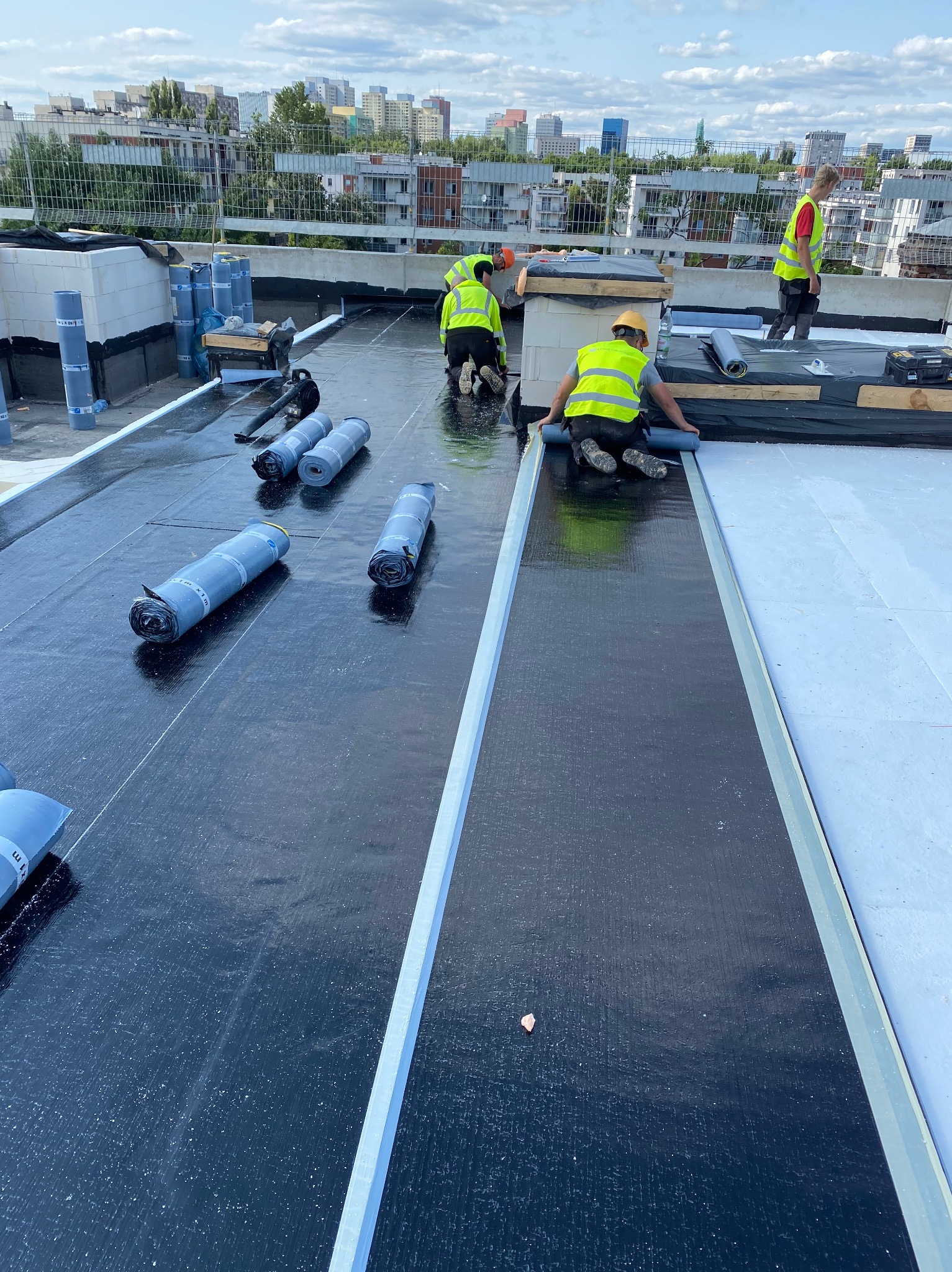 Ekipa w kamizelkach układa papę na dachu budynku. Widoczne rolki papy, białe płyty izolacyjne i panorama miasta w tle. Praca na wysokości.