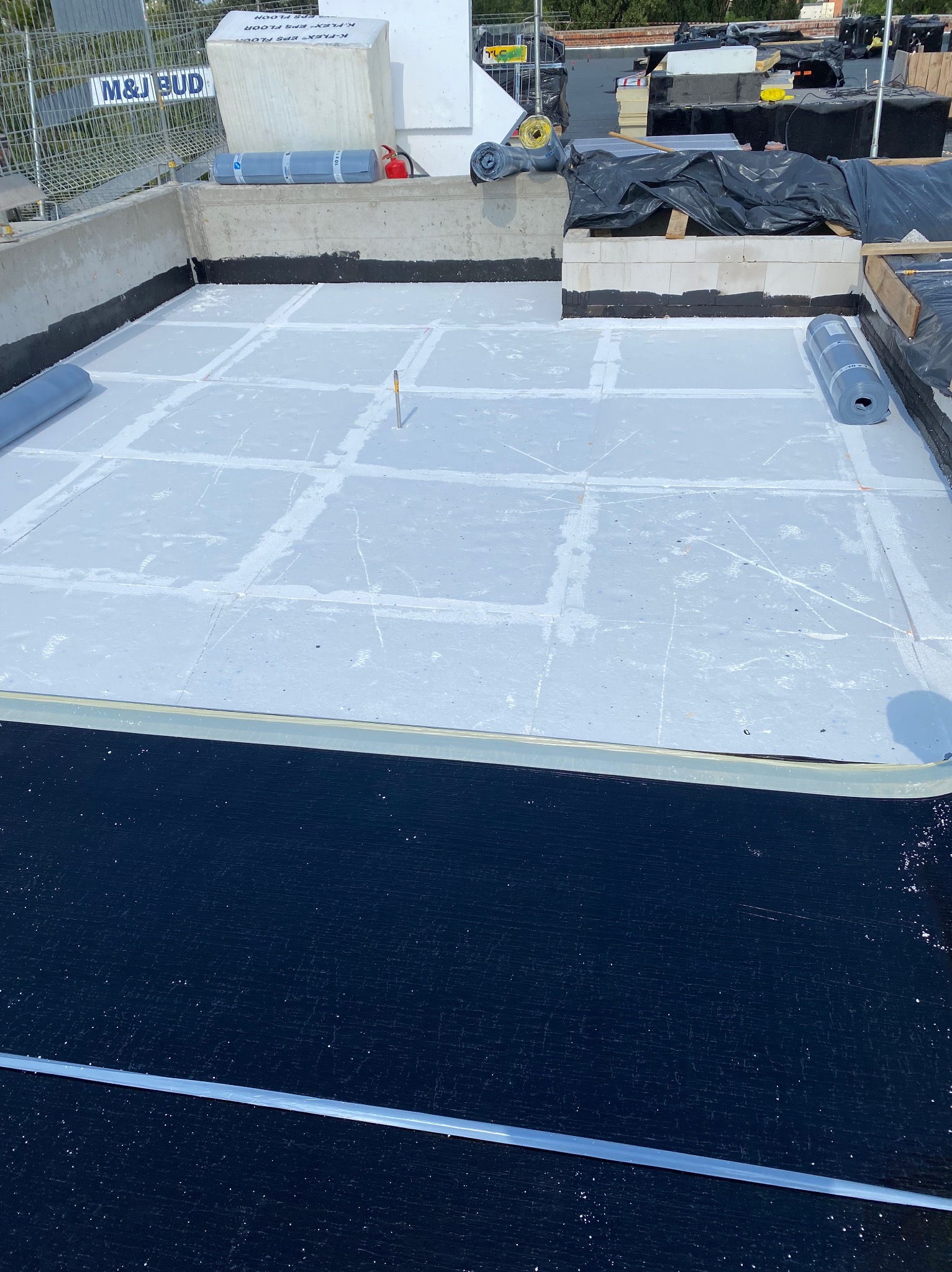 Białe płyty izolacyjne na dachu budynku, częściowo pokryte czarną hydroizolacją. Rolki materiału izolacyjnego w tle. Prace dekarskie w toku.