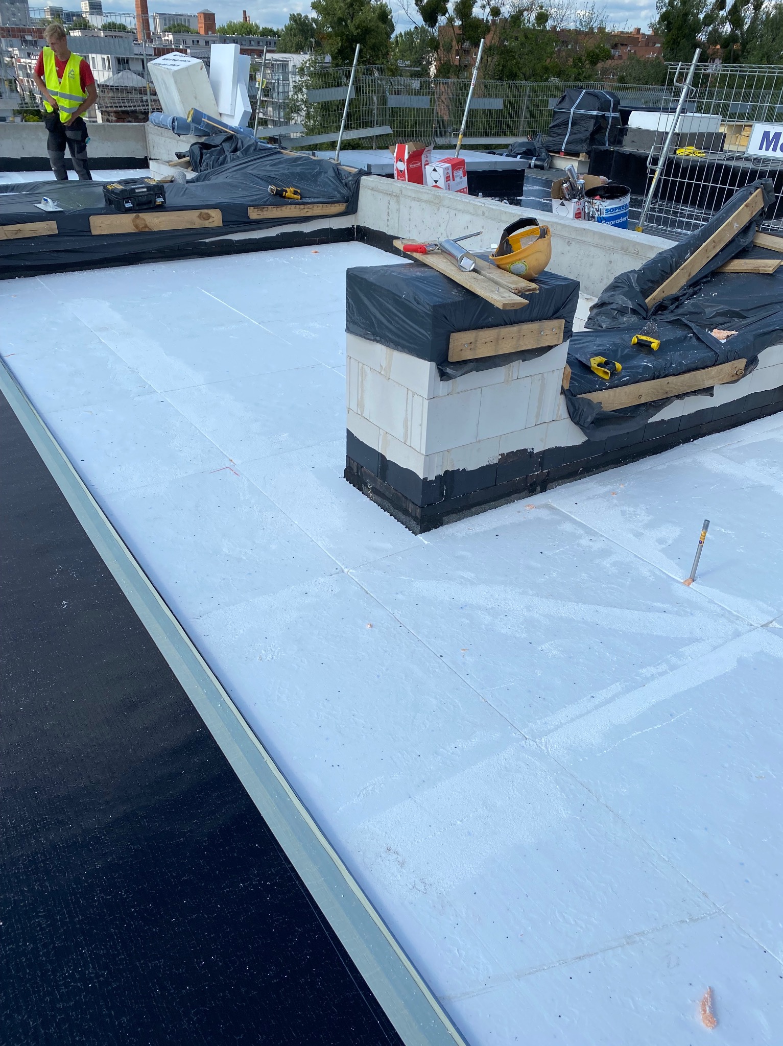 Białe płyty termoizolacyjne na płaskim dachu w trakcie montażu. Widoczne materiały budowlane i narzędzia, pracownik w oddali. Etap prac przed ostatecznym wykończeniem.