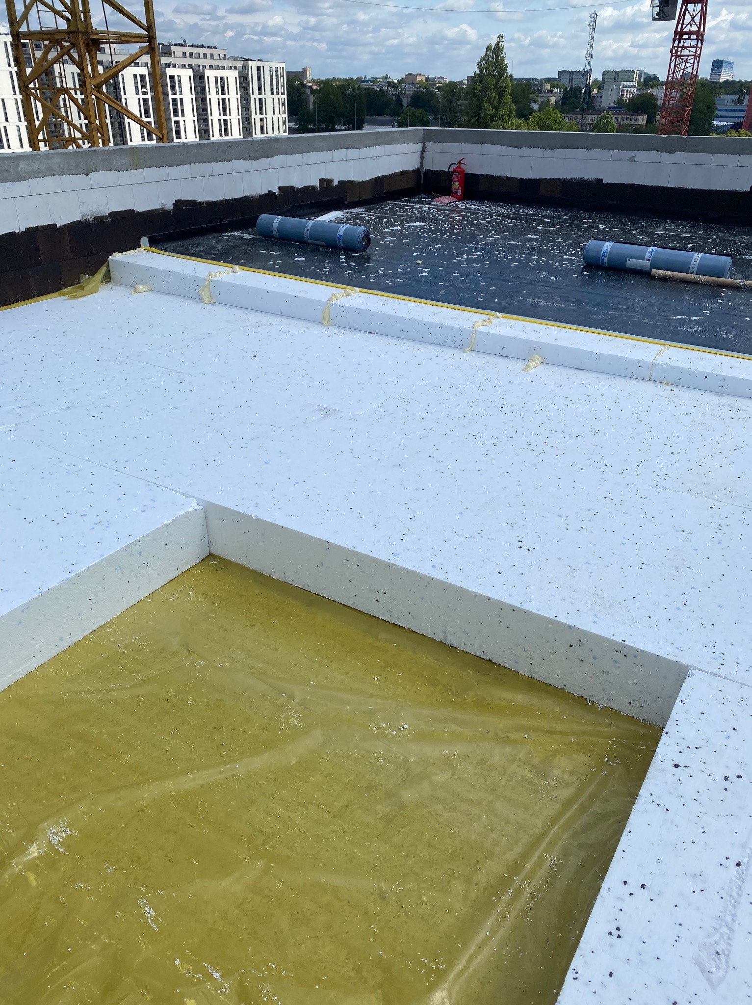 Ocieplenie płaskiego dachu styropianem EPS 100, zabezpieczonego folią hydroizolacyjną. Widoczne rolki papy i gaśnica. W tle budynki i żurawie budowlane.