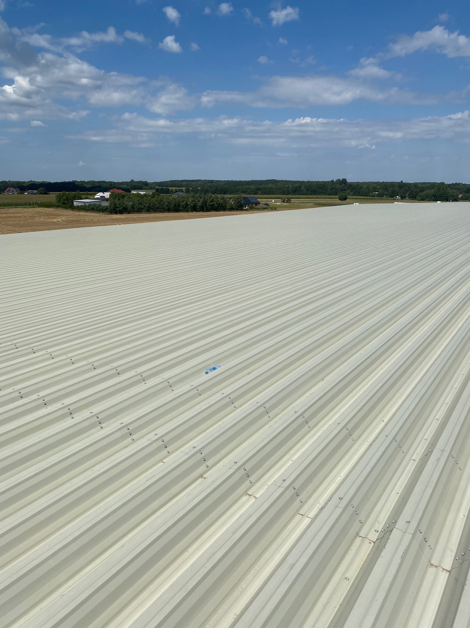 Szeroki, blaszany dach z wyraźnymi liniami w Zduńskiej Woli. Widok z góry, w tle błękitne niebo z chmurami i fragment krajobrazu. Nowoczesne pokrycie.