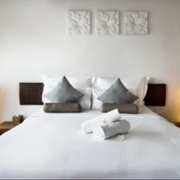 Biała pościel hotelowa z ręcznikami i kwiatami, drewniane lampy podłogowe i dekoracje ścienne z motywem kwiatowym, tworzące przytulną i czystą atmosferę.