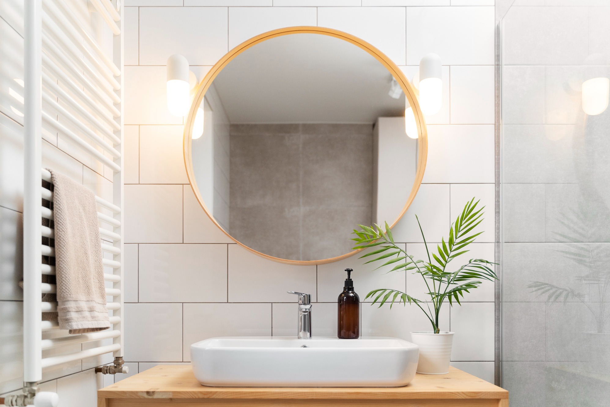 Jasna łazienka z okrągłym lustrem, białą umywalką, grzejnikiem drabinkowym z ręcznikiem i rośliną doniczkową. Świeże, minimalistyczne wnętrze po remoncie.