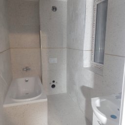 Mykola Dushuk - Jasna łazienka z wanną, umywalką i oknem, wykończona płytkami w drobny wzór. Widoczne punkty instalacji wodnej i elementy armatury.