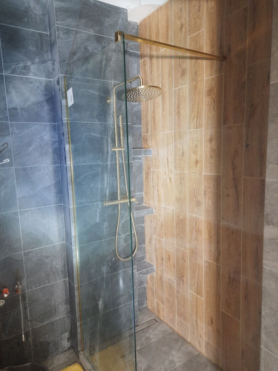 Nowoczesna kabina prysznicowa z deszczownicą w odcieniach szarości i drewna, oddzielona szklaną ścianką. Widoczne półki wkomponowane w ścianę.