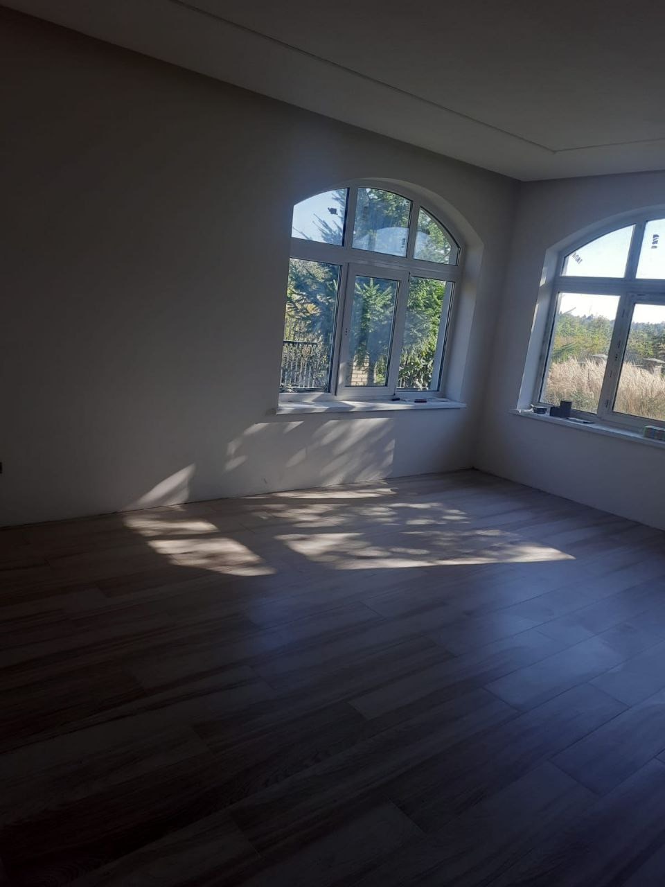 Jasne wnętrze po remoncie z parkietem i dwoma oknami łukowymi, wpadające światło słoneczne tworzy cienie na podłodze. Minimalistyczny wystrój.