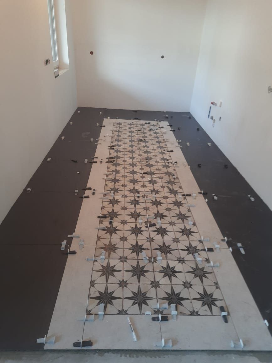 Układanie wzorzystych płytek na podłodze w korytarzu. Widoczne krzyżyki dystansowe i fragmenty ciemnych płytek po bokach. Ujęcie z perspektywy podłogi.