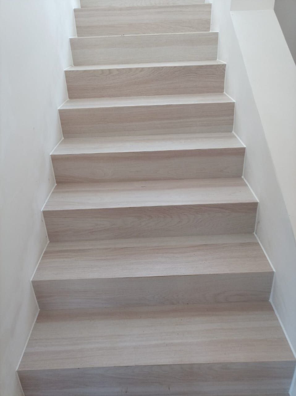 Jasne, drewnopodobne schody z precyzyjnym wykończeniem krawędzi, widoczne z dołu. Minimalistyczny design z białymi ścianami.