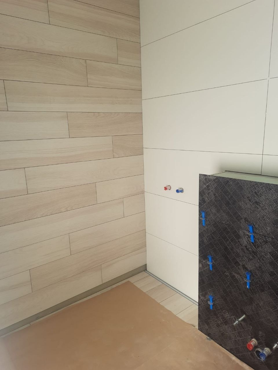 Ściana z płytek imitujących drewno i białe płytki w łazience w Warszawie. Widoczne niebieskie krzyżyki glazurnicze i instalacja wodna.