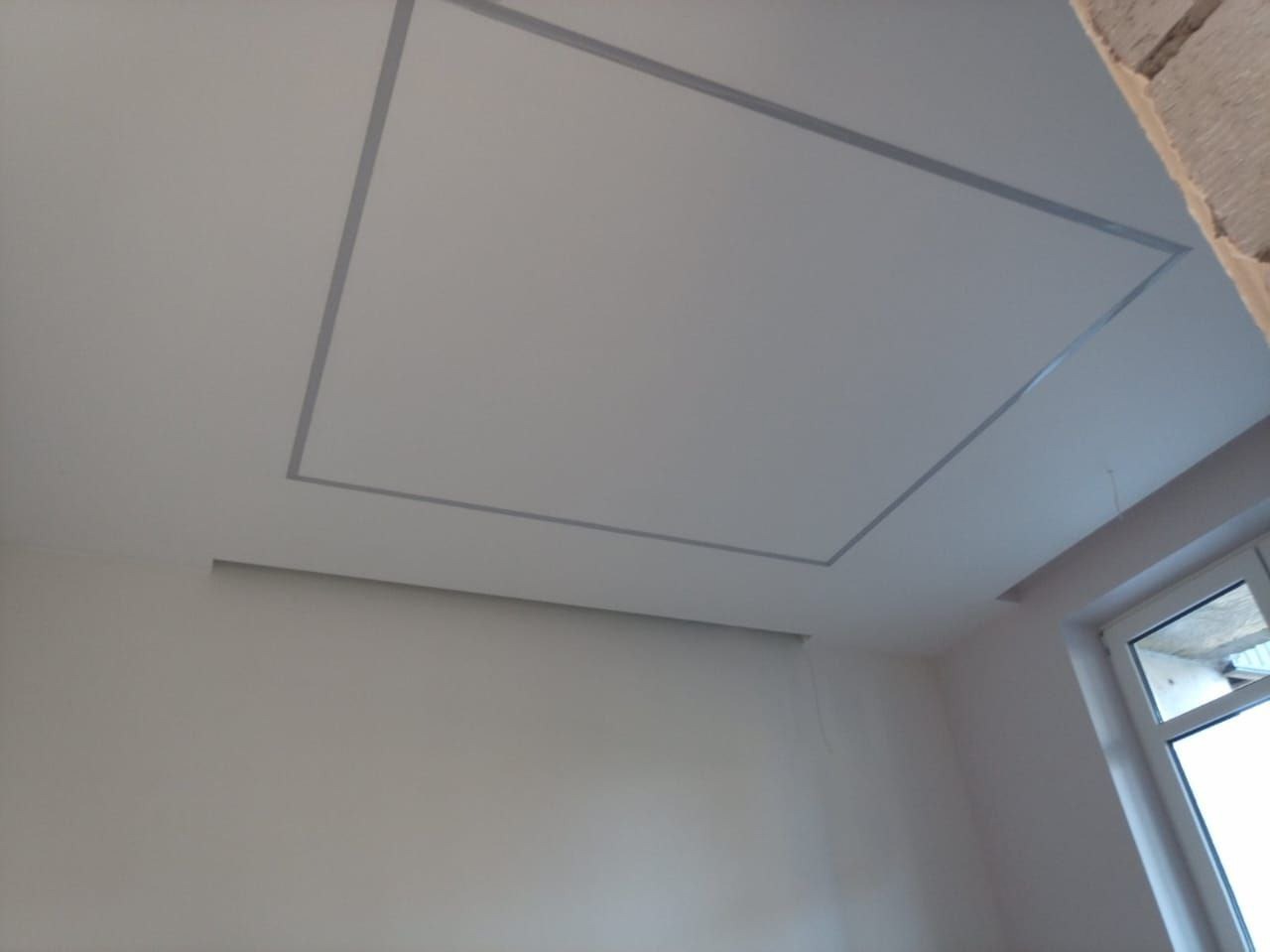 Wykończony sufit podwieszany z szarą ramką, widok z dołu, fragment okna po prawej. Gładkie ściany w jasnym odcieniu beżu.