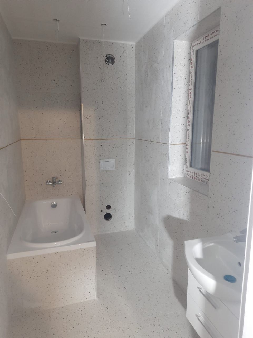 Jasna łazienka z wanną, umywalką i oknem, wykończona płytkami w drobny wzór. Widoczne punkty instalacji wodnej i elementy armatury.