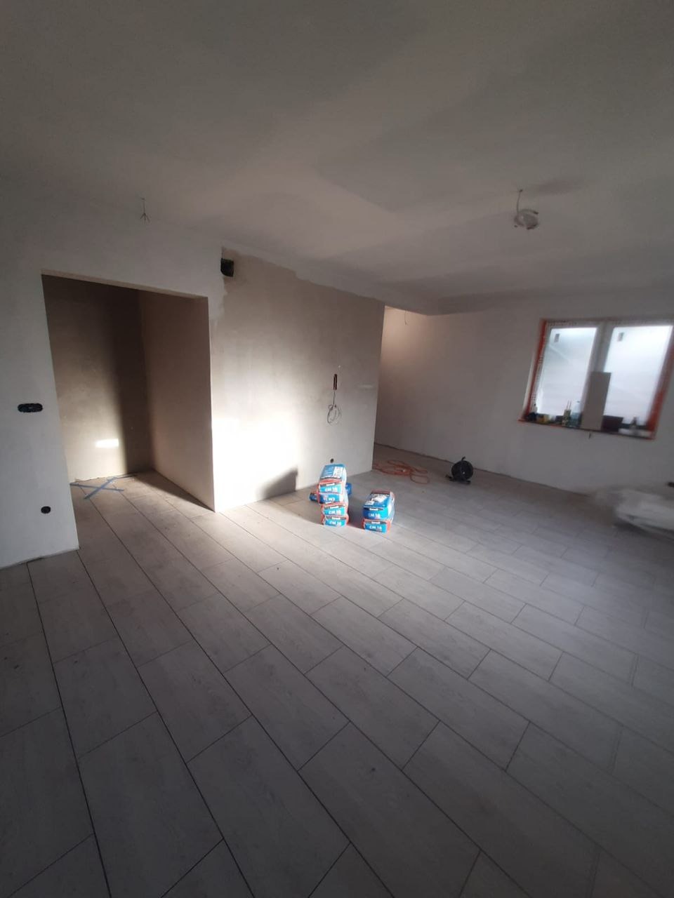 Układanie podłogi z płytek imitujących drewno w jasnym, surowym wnętrzu. Na posadzce leżą worki z zaprawą. Widoczne przygotowane ściany i okno.