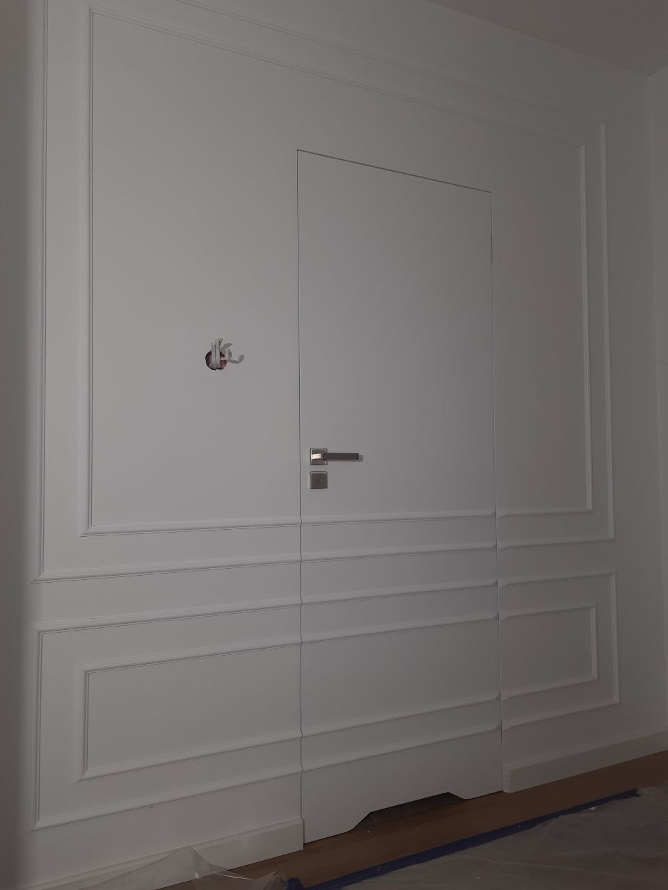 Biała ściana z ukrytymi drzwiami i ozdobnymi panelami. Widoczny fragment podłogi z zabezpieczeniem malarskim. Minimalistyczny design.