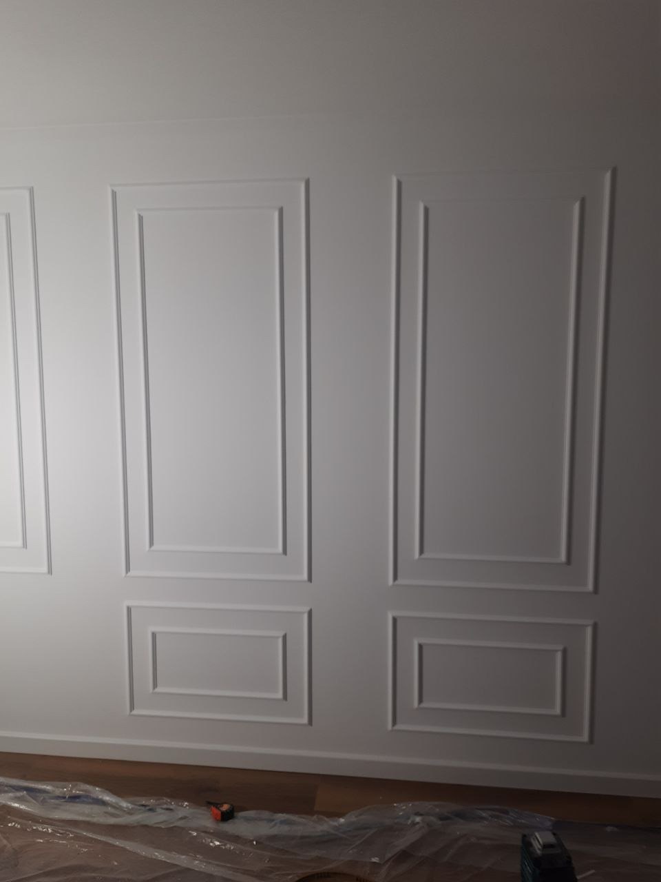 Biała ściana z dekoracyjnymi, symetrycznymi panelami w stylu klasycznym. Na podłodze folia ochronna. Ukończona aranżacja wnętrza.