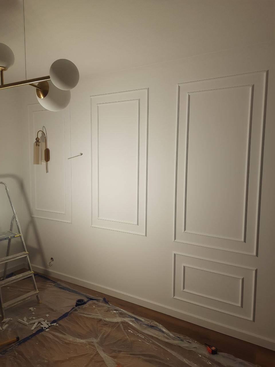 Biała ściana z dekoracyjnymi ramkami, oświetlona nowoczesną lampą. Widoczna drabina i folia ochronna na podłodze, sugerujące trwające prace wykończeniowe.