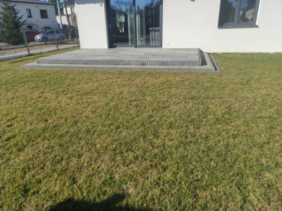 Nowoczesny taras z szarej kostki brukowej przy białym domu z dużymi oknami, otoczony trawnikiem. Widok z poziomu trawnika.