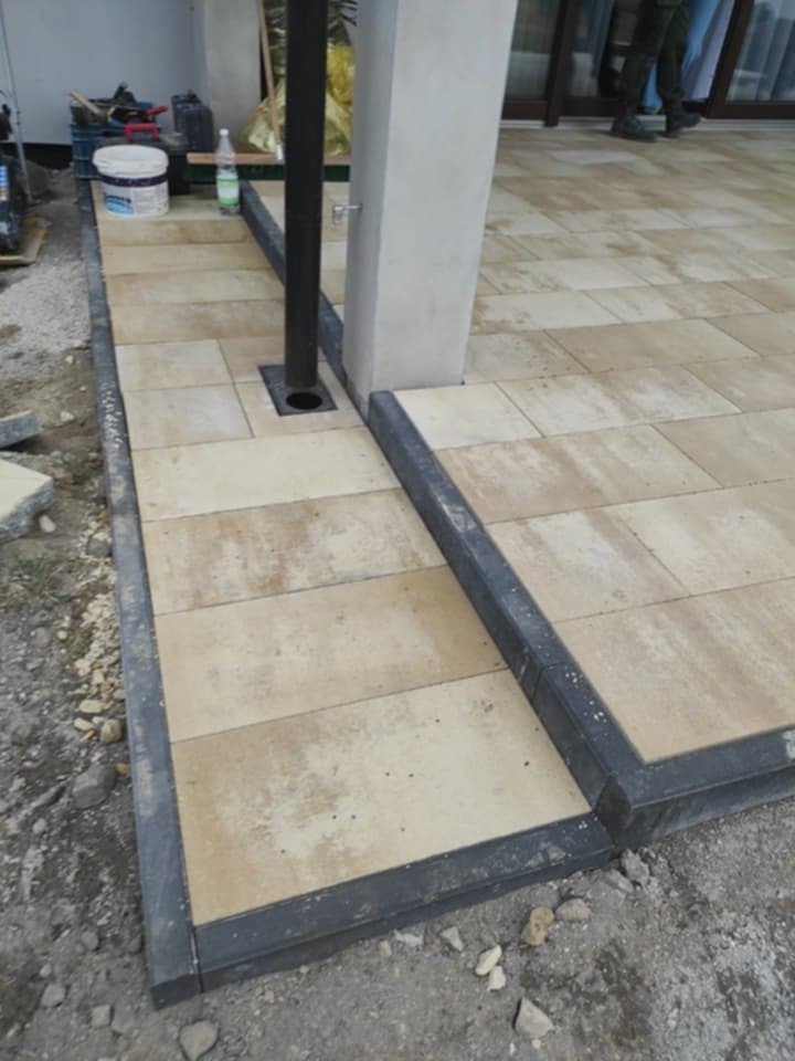 Beżowa kostka brukowa ułożona wokół filaru i rury odpływowej, obramowana ciemnoszarym obrzeżem. Widoczne narzędzia i materiały budowlane w tle.