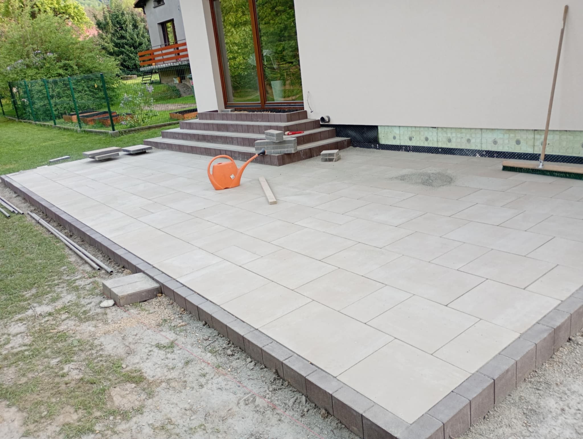 Jasna kostka brukowa na tarasie z obrzeżami, widok na schody i dom. Pomarańczowa konewka i szczotka do zamiatania. Częściowo ułożona kostka przy schodach.