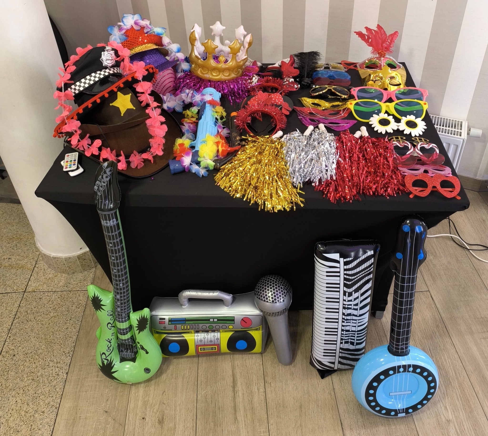 Akcesoria do fotobudki: kapelusze, korony, okulary, peruki, pompony, dmuchane gitary i mikrofon na stole z czarnym obrusem. Różnorodność i kolorowe rekwizyty.
