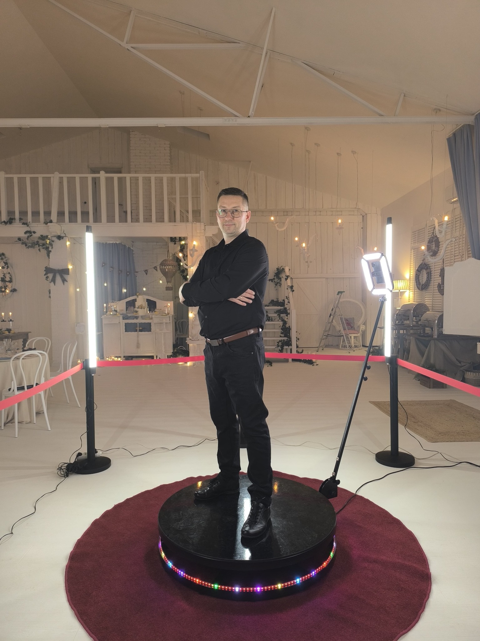 Mężczyzna w czarnym ubraniu stoi na platformie 360 z oświetleniem LED w udekorowanym wnętrzu, prawdopodobnie podczas imprezy.