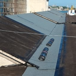 Usługi Ogólno Budowlane Dekarskie - Fragment płaskiego dachu pokrytego nową papą termozgrzewalną w kolorze szarym, z widocznymi rolkami papy. W tle budynki przemysłowe i błękitne niebo.