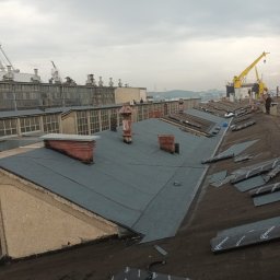 Usługi Ogólno Budowlane Dekarskie - Wykończony fragment dachu z papy termozgrzewalnej w Lubichowie, z widocznymi kominami i częściowo rozłożonymi rolkami papy. W tle budynki przemysłowe i dźwigi.