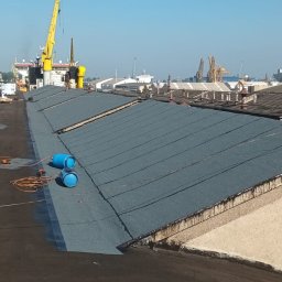 Usługi Ogólno Budowlane Dekarskie - Prace dekarskie na rozległym dachu przemysłowym. Widoczne rolki papy, butle gazowe i częściowo pokryta powierzchnia. W tle dźwigi portowe i zabudowa miejska.