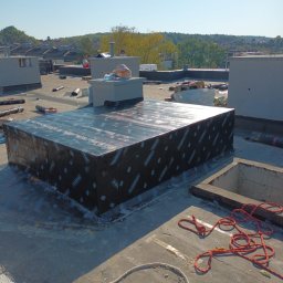 Usługi Ogólno Budowlane Dekarskie - Prace na dachu płaskim: izolacja komina wentylacyjnego czarną membraną. W tle widoczni pracownicy, inne kominy i elementy dachu. Słoneczny dzień.