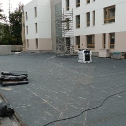Usługi Ogólno Budowlane Dekarskie - Wykończony, płaski dach budynku pokryty papą termozgrzewalną. Zrolowane arkusze papy leżą na powierzchni. W tle rusztowanie i elewacja budynku.