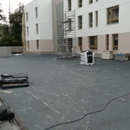 Wykończony, płaski dach budynku pokryty papą termozgrzewalną. Zrolowane arkusze papy leżą na powierzchni. W tle rusztowanie i elewacja budynku.