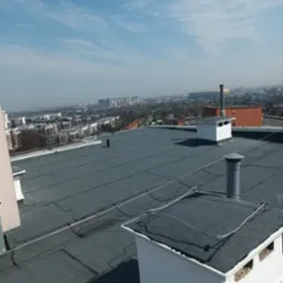 Szary, płaski dach pokryty papą termozgrzewalną z kominami wentylacyjnymi, zabezpieczony odgromowo. W tle zabudowa miejska i błękitne niebo.